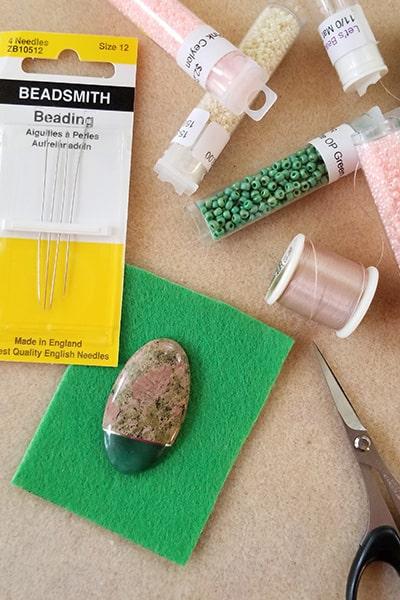 Stone cabochon on backing with items needed for beadweaving: needles, beads, thread, and scissors.